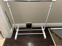White and chrome expandable wardrobe frame shown against wall on dark wood floor, visible base, vertical supports, and chrome hanging bar.