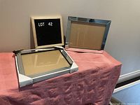 Two mirrored photo frames on a pink fabric surface, one upright, one flat with corner guards, with a small black letter board displaying 'LOT 42' in the background.