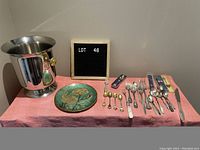 Full view of the lot including ice bucket, brass enameled plate, assorted spoons and cutlery, and boxed cutlery sets arranged on pink cloth.