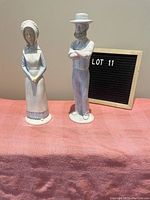 Pair of Grandma and Grandpa ceramic figurines standing side by side on a pink cloth background with a small letter board in the background showing 'LOT 11'.