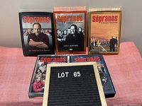 Five DVD boxed sets of The Sopranos seasons one to five, arranged on a pink surface.