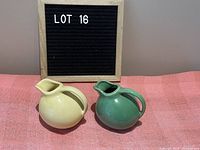Two vintage creamers, yellow and green color, placed on pink cloth with a letter board in background