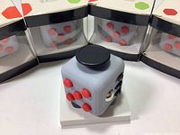 Front and angled view of two new fidget cubes and four boxed fidget cubes in the background showing red buttons and black interactive parts on light gray cubes.