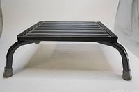 Front view of black bariatric step stool showing metal platform with parallel grooves and rubber-capped legs.