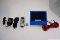 Photo of the blue-framed 7 inch monitor, power adapter, remote control with batteries, and various cables including red and black wires.