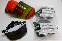 Orange Salisbury hard hat with clear arc flash protection face shield attached, headgear adjustment mechanism visible, placed next to three sealed black V-Gard accessory headgear units.