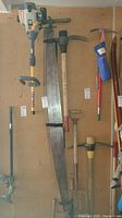 Full view of the antique two man hand saw hanging on a peg board with other tools around it, showing its long curved blade and wooden handle.