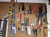 Wide view of pegboard with hand tools mounted including saw, pipe wrench, scrapers, levels, garden trowels, clamps, and other small tools.