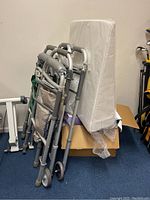 Three folding walkers made of aluminum with padded arm grips and multiple fabric storage bags folded upright leaning against a cardboard and wall, with wedge pillow behind.