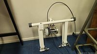 Folded white metal frame of indoor bike trainer on floor with visible cable attached.