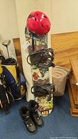 Full view of kids snowboard with Spider-Man graphics, red helmet atop, and black boots on floor.