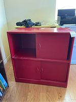 Red wooden cabinet with two sliding doors on the bottom and one on the top, missing one door, with visible wear and some dust.