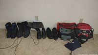Photo showing three pairs of men's boots, one pair of shoes, and three small backpacks grouped on carpeted floor against a wall