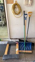 Photo of snow shovels, push broom, garden tool, and extension cord hanging on wall and leaning on floor, showing overall condition and setup.