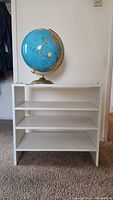 Front view of small white 3-tier bookshelf with blue desktop globe placed on top against wall.