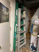 Two step ladders leaning against a wall near a door and water heater, stacked together, one green-sided and one fully aluminum.