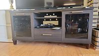 Front view of dark wood TV stand with glass cabinet doors, central drawer, and open shelving, displaying size and design.