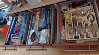 Right two drawers showing stainless steel flatware organized in trays and additional kitchen tools and utensils in the adjacent drawer.
