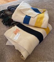 Four folded vintage wool blankets stacked on carpet, showing Hudson's Bay striped blanket on top and plaid and other blankets underneath.