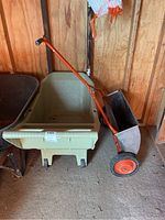 Plastic wheelbarrow and metal seed spreader side by side inside a wooden shed, showing the wheelbarrow’s one front wheel and the seed spreader’s orange handle and wheels.