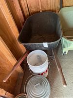 Full view of metal wheelbarrow with black tub and wooden handles in shed, with white bucket and metal lid underneath.