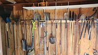 Wide view showing assortment of long-handled yard tools hung on wooden rack in shed including shovels, post hole diggers, and digging forks.