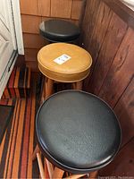 Three bar stools: one gold vinyl seat with wood legs, two black vinyl seats with wood legs, shown together on a carpeted floor near a corner wall.