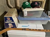 Photo showing collection atop household washer/dryer: two baseball caps (Boston Celtics and Arizona), plastic bins, wooden hockey stick, NFL helmet snack bowl box.