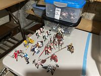 Twelve knight figures and four horses arranged on a white table in front of two stacked bins