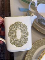 Close-up photo of a square-shaped creamer or small pitcher showing olive green and gold medallion design on white porcelain