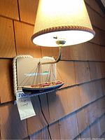 Front view of the ship lamp mounted on a wooden shingle wall, showing beige fabric lampshade and detailed model sailboat base with rope border.