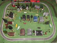 Wide view of the model train village including buildings, vehicles, trees, and oval grey track surrounding scene.