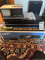 Lot overview showing Samsung DVD Recorder & VCR in box, Toshiba VHS/DVD player, Emerson TV, alarm clock radio and a Sony speaker.