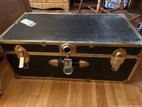 Full view of the black vintage trunk with brass corner reinforcements and latches on a wood floor.