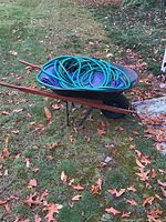 Side angled view of metal wheelbarrow with wooden handles holding a coiled green garden hose and folded blue tarp.