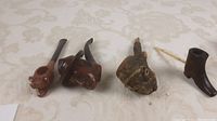 Four carved pipes on fabric backdrop including two figure head pipes, one bark root pipe with twig stem, and one boot-shaped bowl
