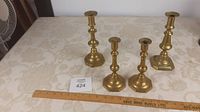 All four brass candle sticks displayed on a surface with a ruler showing size and an inventory tag visible