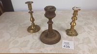 Photo shows three candle sticks standing on a table: one wooden turned candle stick in the center and two brass candle sticks on either side, visible age and wear on all items.