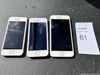 Front view of three used iPhones with powered off screens, placed side by side on dark surface with lot tag visible.