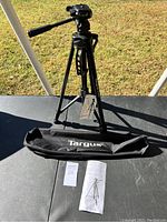 Full view of the Targus camera stand tripod set up outside on a black surface, including the tripod with extended legs, Targus branded carrying bag, and instruction manual.