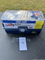 Top front view of rectangular blue tackle box with clear plastic lid showing various fishing gear inside including hooks, floats, tins of swivels and weights.
