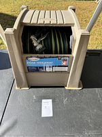 Front view of the beige Suncast Hose Hideaway housing unit with green and yellow coiled garden hose and metal spray nozzle inside.