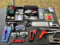 Overview of tools including power drill, measuring tapes, clamps, staplers, goggles, and other accessories on black table.