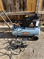 Full side view of blue and black McGraw air compressor showing wheels, handle, and attached hoses on ground