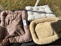 Three new pet beds displayed outdoors: brown fabric cushion bed, white fluffy cushion bed, tan sherpa fleece bed with raised edges.
