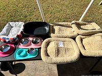 Photo showing four beige plush rectangular pet beds, one large black plastic pet bed, a patterned cushioned pet bed, and various colored feeding bowls and feeders on a table.