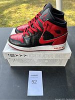Side view of Nike Air Jordan 1 sneakers in black and red atop the original box labeled 'COLOR ME'. Nike swoosh and laces are red, with black leather panels.