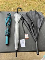 Three umbrellas placed on a black table outdoors: one black umbrella with floral cutouts near the edge, one clear plastic umbrella with black handle and tip, and one folded teal and black striped umbrella with a wrist strap.