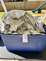 Large silver car tarp folded inside a blue plastic bin, displayed outdoors on a patio with a backyard fence and playground slide in the background.