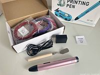 3D printing pen in rose gold color with USB charging cable, USB power adapter, wooden handle tool with metal spatula tip, and filament strands in red and purple colors in packaging.